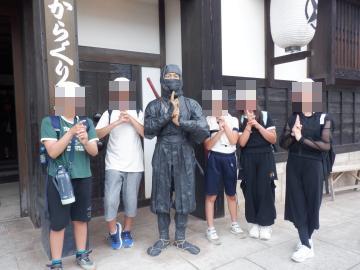 古い町家の前で忍者の恰好をした人物と生徒たちグループが両手を合わせて印を結んで並んでいる