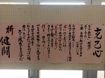 窓の上に掲示されたピンクの横長の紙に黒い筆文字で「克己心」を含む長文の和文が並んだ掲示物