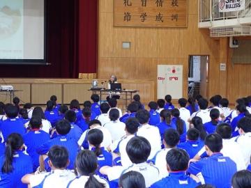 体育館で多数の生徒が床に座って講師の話を聞いている様子