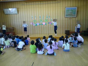 遊戯室で床に座った子どもたちが舞台に立つ数人の子どもの発表を見ている