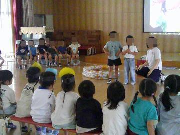 遊戯室の舞台で話す児童と椅子に座ってそれを聞く3，4歳児たち