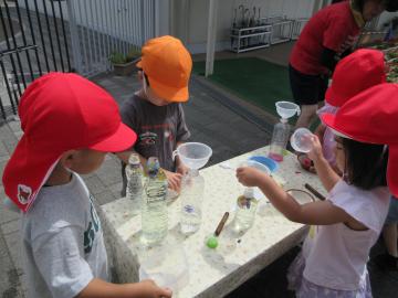 3歳児と4歳児が屋外のテーブルでペットボトルやカップを使って色水遊びをしている様子