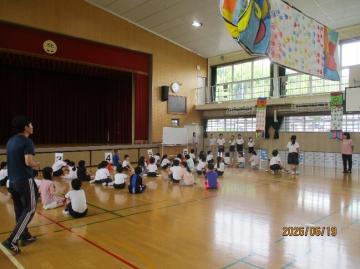 体育館の床に座っている児童たちと前方で指示する教員や舞台と窓から差し込む明るい光が見える風景