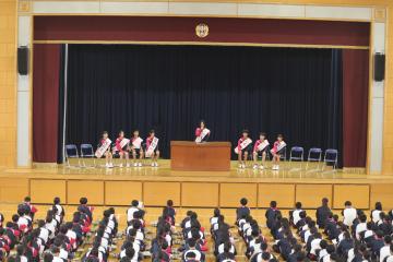 体育館の舞台上に並ぶ候補者と体育館の床に全校生徒が座っている様子1