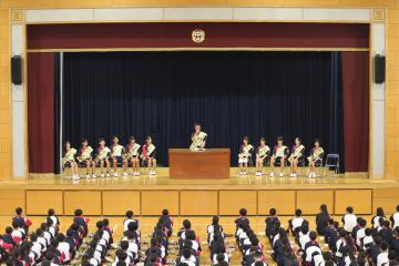 体育館の舞台の演台に立つ生徒と周りに座る生徒とそれを座って見る全校生徒