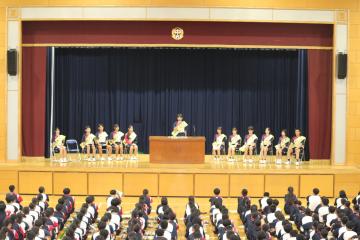 体育館の舞台の演台に立つ生徒と周りに座る候補者と体育館に座って演説を見る全校生徒