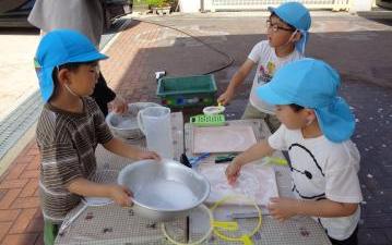 青い帽子をかぶった園児たちが屋外の机で水や器を使って遊んでいる様子