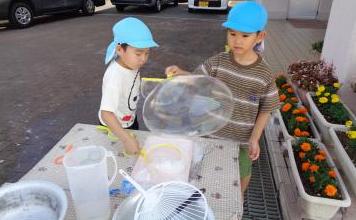青いキャップをかぶった二人の園児がテーブルで大きなシャボン玉を作っている様子。テーブルにトレイなどが並んでいる様子