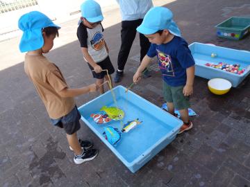 園児たちが屋外の床に置かれた水の入ったプラスチック容器の周りでおもちゃの魚を釣り竿で釣る遊びをしている様子