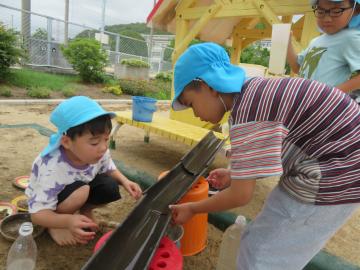 青い帽子をかぶった数人の園児が屋外の遊具のそばで水やおもちゃを使って遊んでいる様子
