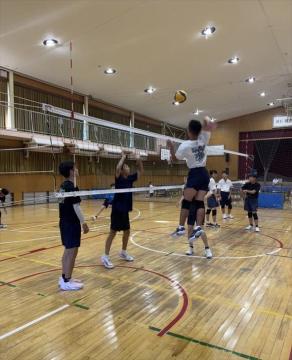 屋内の体育館で数人の児童がネットを挟んでバレーボールの練習をしており一人がジャンプしてボールを打とうとしている場面