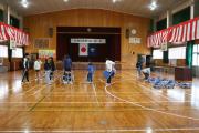 体育館の舞台に日の丸の国旗が掲げられ前方に間隔を空けて並んだ椅子と立つ人々が見える校内の集会の様子