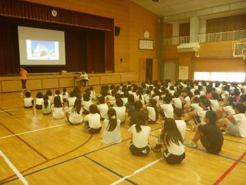 体育館で床に座って投影されたスライドを見ながら説明を聞く児童
