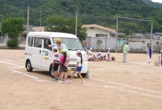 運動場で白い軽バンのそばで大人が道路の渡り方を教えている
