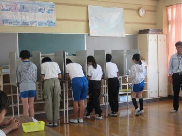 教室の中で仕切りのある投票ブースに並んで記入する制服姿の生徒数名とそばで立つ大人
