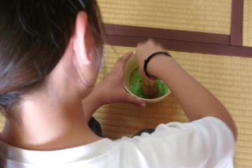 和室で、畳の上に座り茶碗を両手で支え茶筅で抹茶を点てている児童の後ろ姿