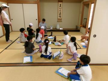 畳の部屋で先生に見守られながら座って公民館長さんの話を聞く児童たちの様子