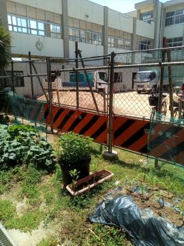 工事用の黒とオレンジの斜め縞の仮囲いとフェンス越しに校舎のような建物と作業車が見え手前に植木鉢や雑草が茂る植栽スペースがある風景