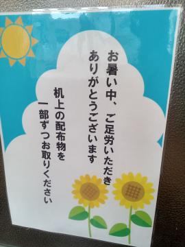 青い背景に太陽とひまわりのイラストと暑い中ご足労いただきありがとうございます机上の配布物を一部ずつお取りくださいと書かれた案内表示