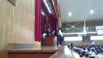 体育館の舞台で数名が話しているのを多数の生徒が座って見ている