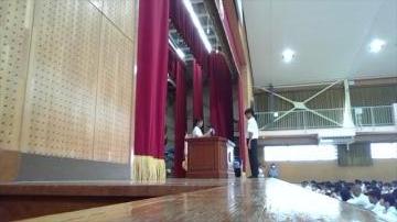 学校の体育館の舞台でカーテンの前に立つ数人と演台に立つ人を低い位置から舞台端と観客席を含めて見渡せる視点で捉えた写真