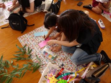 室内で、親子で床に置かれた短冊に願い事をかいたり笹飾りを作っている様子