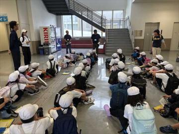 ロビーに座った児童たちの前に立つスタッフから見学前の施設の話を聞いている