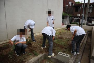 5人の生徒が鎌やスコップを持ち、協力しながら校門付近の花壇の雑草を抜いています。