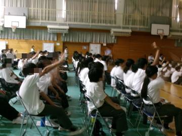 体育館の座席に並んで座る生徒たちが手を挙げています。