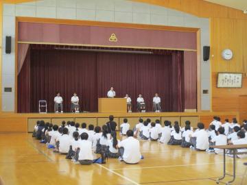 体育館の舞台で数人の生徒が並び前方の床に座っている多くの生徒が発表を聞いている