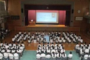 体育館で制服を着た多数の生徒が座ってスクリーンと演壇のある舞台に向かって集会に参加している光景