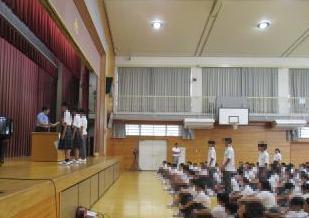 体育館の舞台上に数名の生徒と教員が立ち床に座って整列する多くの生徒たちが前を向いている集会の光景