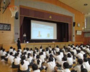 体育館のステージ上のスクリーンで資料が映されている前で床に並んで座り発表を聞く白い服の生徒たちと壇上の生徒