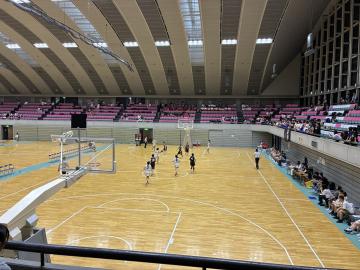 屋内の体育館で観客席を背にバスケットボールの試合が行われているコート上に選手と審判がいる