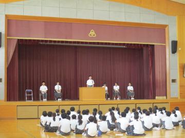 体育館の舞台で演壇に立つ人と左右に座る数人を前に床に座っている生徒たちが行事を聞いている光景