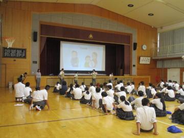 体育館の舞台前に座ってスクリーンを見ている制服の生徒たちと前で説明している数名の大人と数名の生徒