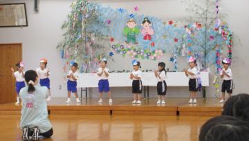 七夕の飾りが施された壁の前の舞台に並んだ子どもたちが先生や保護者を前に歌や発表をしている