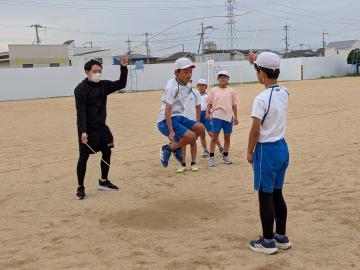 校庭でスポーツ協会の指導者と児童が持った長縄を回し数人の児童が順番にジャンプしている様子