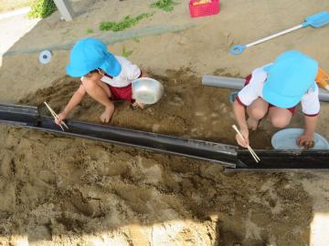 子ども二人が砂場にしゃがんで、樋に流れてくる物を箸でつかみながら遊んでいる