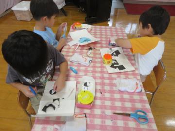 三人の子どもがテーブルで紙やはさみやのりを使って工作をしている様子