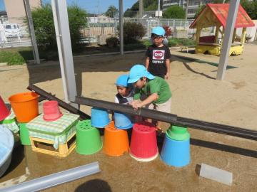 子どもたちがプラスチックのバケツや筒を並べて水を流す道を作っている様子
