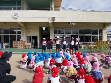 幼稚園の園庭で園児たちが座って中学生がその前で発表しているところ