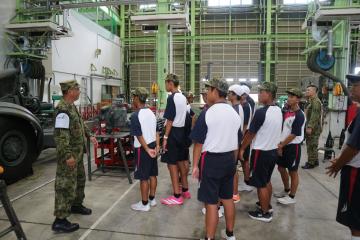 自衛隊の屋内の産業設備の中で学生たちが迷彩服の指導者から説明を受けている