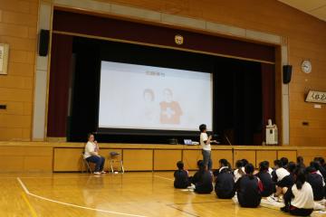 体育館の舞台のスクリーンに投影されたスライドを漫才師2人が説明する様子