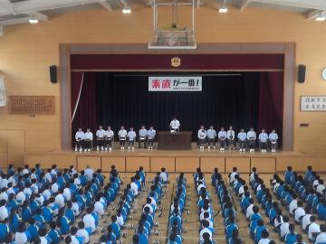 体育館の舞台に立つ生徒と床に整列して座る多くの生徒がいる集会