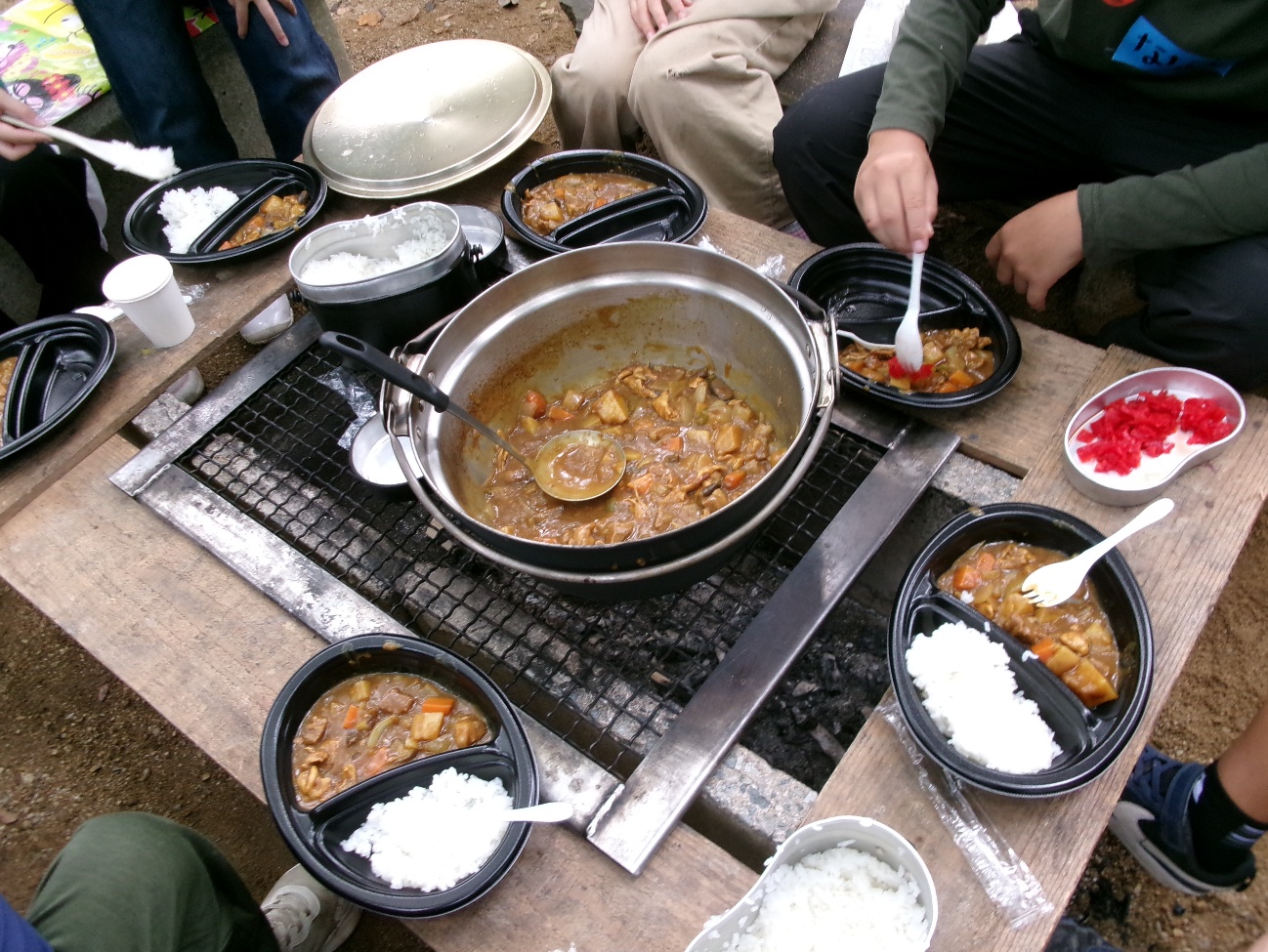 テーブルの中央に鍋に入ったカレーが置かれ周囲にご飯とカレーが盛られた複数の皿が並び人がスプーンでよそう写真