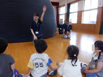 遊戯室の床に座る子どもたちを前にして片手を挙げて説明するスタッフと部屋の奥に置かれた大きな黒いドーム型装置のプラネタリウム