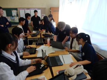 教室で複数の生徒が机を囲んでノートパソコンや資料を使いながら協力して作業している様子