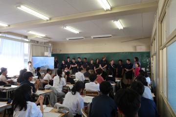教室で前に並んだ学生グループが黒板前で発表しているのを机に座った他の学生たちが聴いている写真