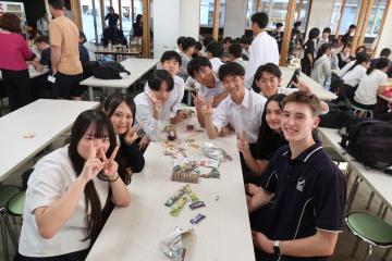 カフェテリアの長いテーブルに集まった学生たちが笑顔でピースサインをしている